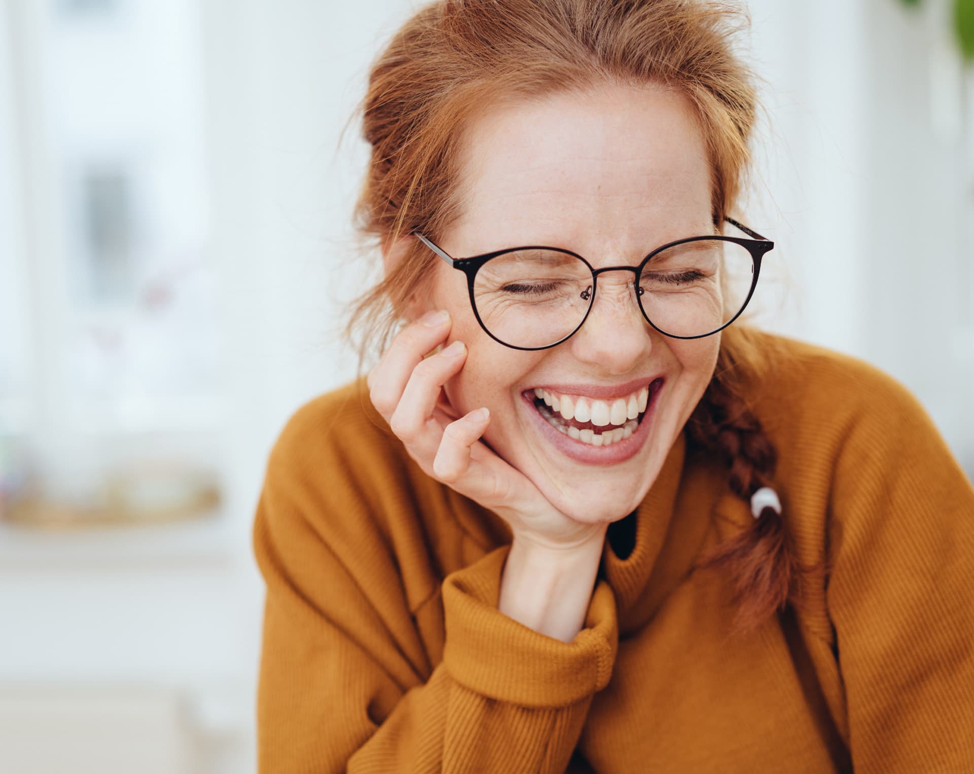 Laughing woman in glasses and an orange jumper, chin resting on her hand